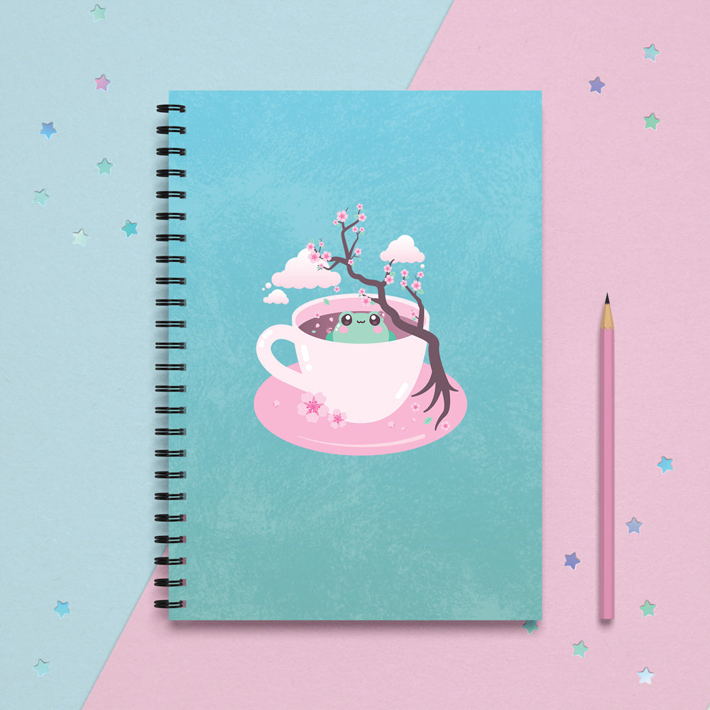 spiral notebook journal planner with a kawaii cute green frog sitting in a pink tea cup with pastel japanese sakura cherry blossom tree and petals with clouds in the sky