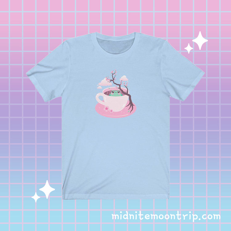 t-shirt with a kawaii cute green grog sitting in a pink tea cup with japanese sakura cherry blossom tree and petals and clouds in the sky