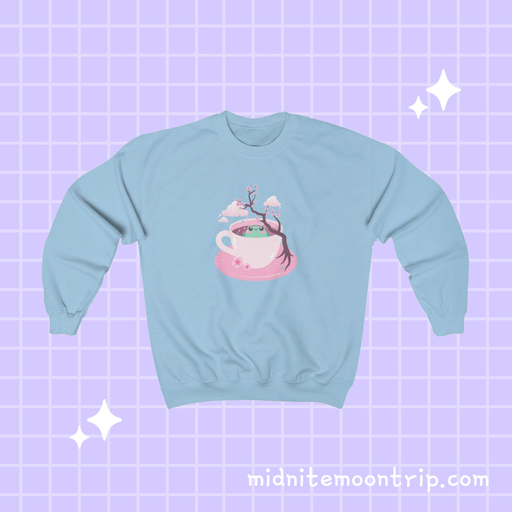 crewneck sweatshirt with a kawaii cute green frog sitting in a pink tea cup with pastel japanese sakura cherry blossom tree and petals with clouds in the sky