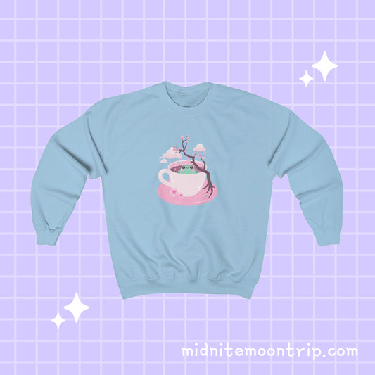 crewneck sweatshirt with a kawaii cute green frog sitting in a pink tea cup with pastel japanese sakura cherry blossom tree and petals with clouds in the sky